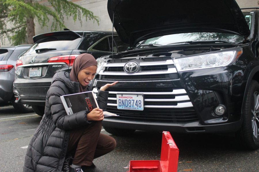 Senior Hebaq Farah crouches in front of her car with a toolbox in front of her and a textbook in hand. Unable to fix her car, Hebaq Farah worries that the things she learned in school are of no help to her in this situation.